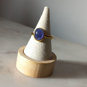 K&ouml;nnte beinhalten: Ein goldener Ring mit einem blauen ovalen Edelstein, der in einer Zargenfassung gefasst ist. Der Ring wird auf einem h&ouml;lzernen Ringst&auml;nder pr&auml;sentiert.