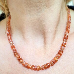 Peut inclure: Un collier compos&eacute; de pierres pr&eacute;cieuses orange &agrave; facettes, entrecoup&eacute;es de petites perles dor&eacute;es. Le collier est port&eacute; autour du cou d'une personne, mettant en valeur la couleur et le design du bijou. Les pierres pr&eacute;cieuses ont un aspect brillant et poli.