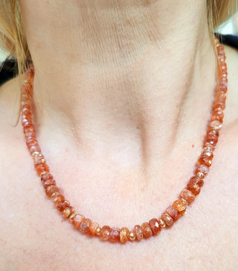 Peut inclure: Un collier compos&eacute; de pierres pr&eacute;cieuses orange &agrave; facettes, entrecoup&eacute;es de petites perles dor&eacute;es. Le collier est port&eacute; autour du cou d'une personne, mettant en valeur la couleur et le design du bijou. Les pierres pr&eacute;cieuses ont un aspect brillant et poli.
