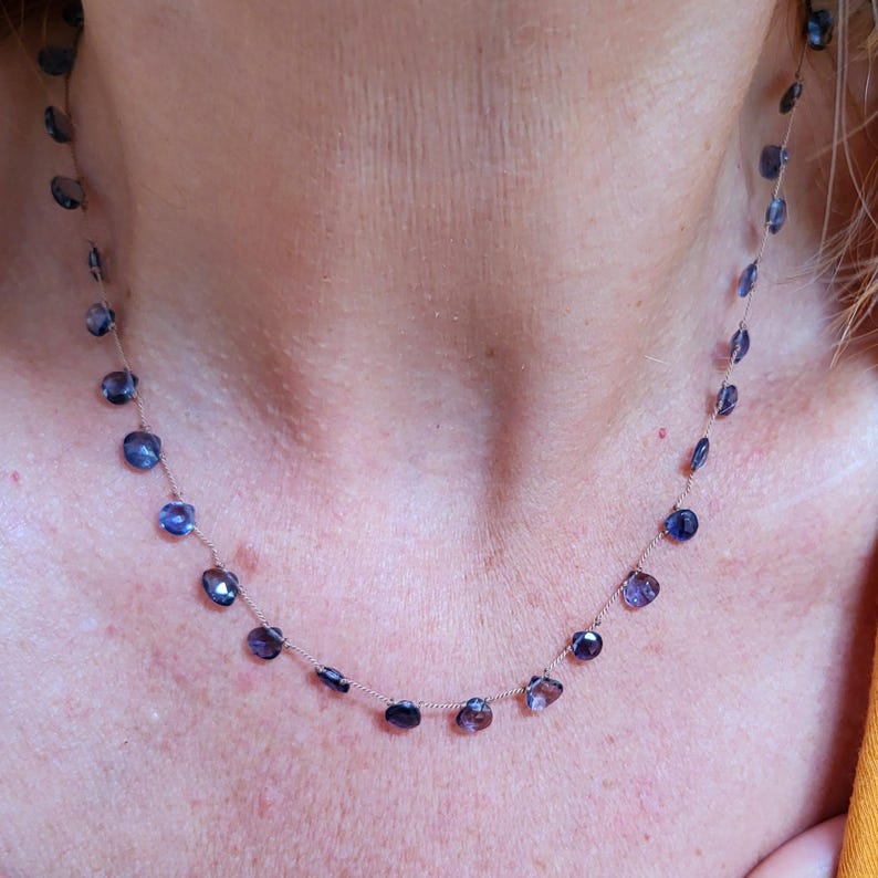 May include: A delicate silver chain necklace with small, round, dark blue gemstones. The necklace is worn by a person, but the focus is on the necklace itself.