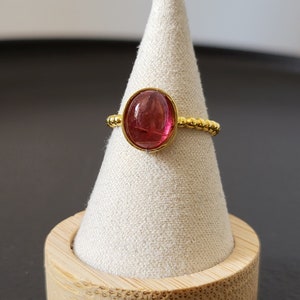 May include: A gold ring with a pink oval gemstone set in a beaded band. The ring is displayed on a small wooden stand.