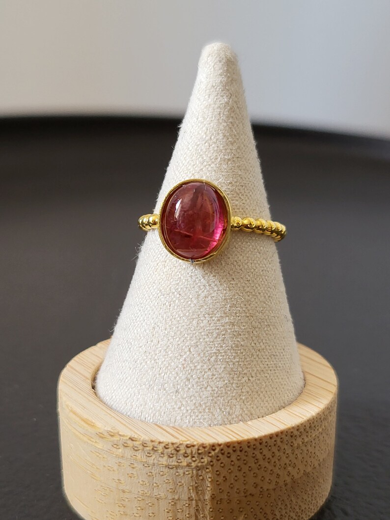 May include: A gold ring with a pink oval gemstone set in a beaded band. The ring is displayed on a small wooden stand.