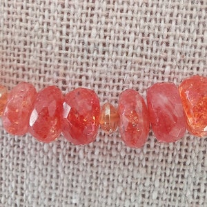 Peut inclure: Gros plan sur un collier orn&eacute; de perles de soleil rouge-orang&eacute; facett&eacute;es. Les perles, de tailles et formes vari&eacute;es, sont s&eacute;par&eacute;es par de petites perles transparentes. Le collier est pr&eacute;sent&eacute; sur une surface textur&eacute;e de couleur neutre.