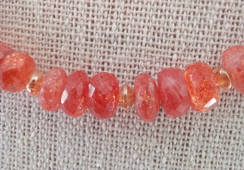 Peut inclure: Gros plan sur un collier orn&eacute; de perles de soleil rouge-orang&eacute; facett&eacute;es. Les perles, de tailles et formes vari&eacute;es, sont s&eacute;par&eacute;es par de petites perles transparentes. Le collier est pr&eacute;sent&eacute; sur une surface textur&eacute;e de couleur neutre.