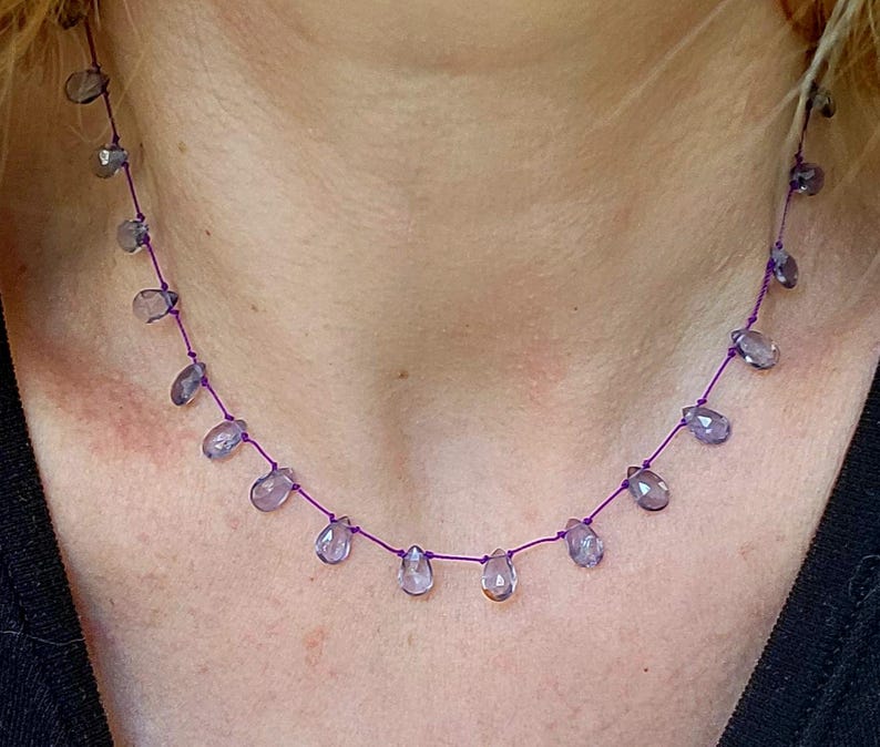 May include: A delicate necklace featuring teardrop-shaped, translucent purple beads strung on a thin, vibrant purple cord. The beads are evenly spaced, creating a minimalist and elegant design. This necklace is a piece of jewelry.
