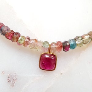 May include: A necklace made of multi-colored gemstone beads with a square-shaped pendant featuring a pink gemstone.
