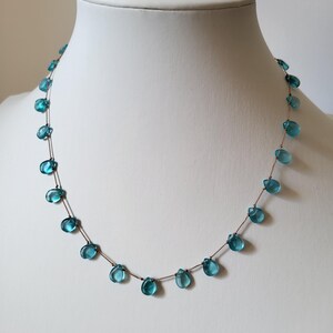 Pu&ograve; includere: Una delicata collana con perline a forma di goccia, di colore azzurro chiaro, infilate su una sottile catena marrone. Le perline sono distanziate in modo uniforme, creando un design minimalista ed elegante. La collana &egrave; esposta su un manichino bianco.