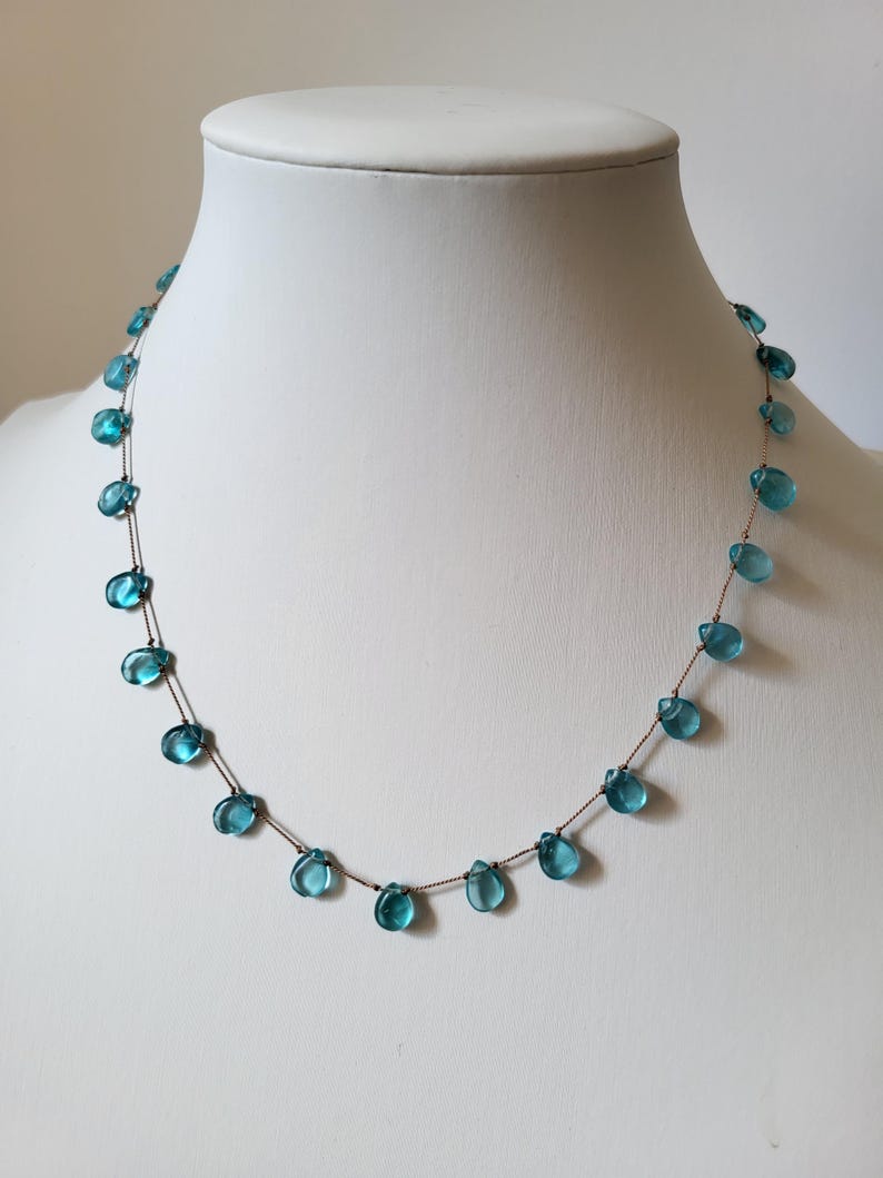 Pu&ograve; includere: Una delicata collana con perline a forma di goccia, di colore azzurro chiaro, infilate su una sottile catena marrone. Le perline sono distanziate in modo uniforme, creando un design minimalista ed elegante. La collana &egrave; esposta su un manichino bianco.