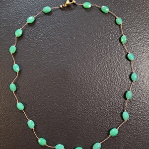 May include: A delicate necklace featuring oval-shaped, light green gemstones strung on a thin, gold-colored chain. The necklace has a gold-colored clasp. The gemstones are evenly spaced along the chain, creating a simple yet elegant design.
