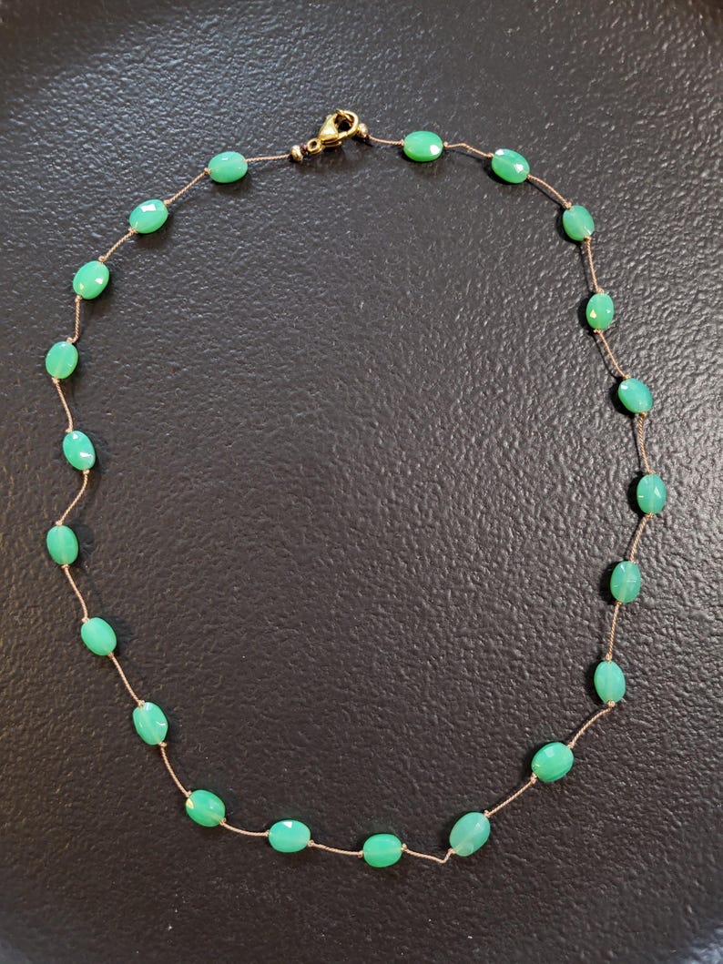 May include: A delicate necklace featuring oval-shaped, light green gemstones strung on a thin, gold-colored chain. The necklace has a gold-colored clasp. The gemstones are evenly spaced along the chain, creating a simple yet elegant design.