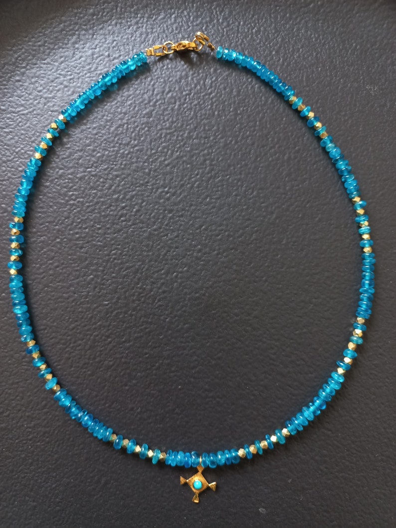 May include: A turquoise blue gemstone necklace with a gold pendant featuring a turquoise stone in the center. The pendant is in the shape of a cross with four points.