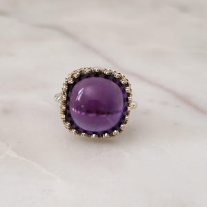 May include: A silver ring featuring a large, round, deep purple amethyst gemstone. The gemstone is set in a decorative, textured bezel. The ring is displayed on a white marble surface.