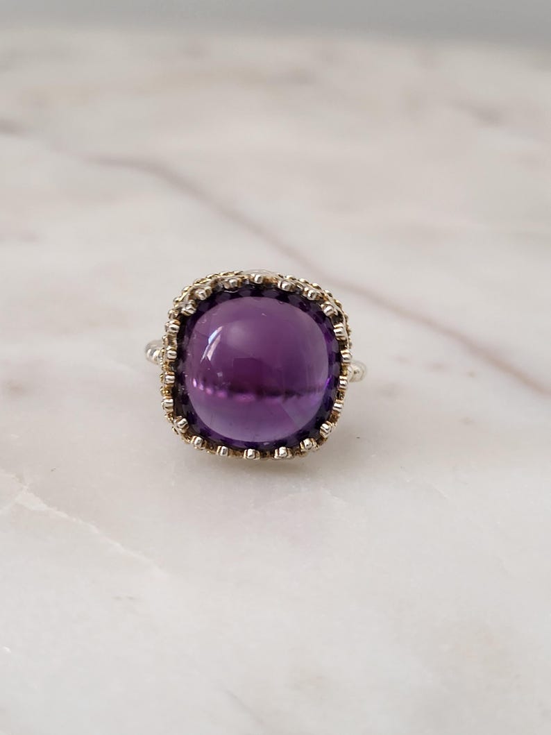 May include: A silver ring featuring a large, round, deep purple amethyst gemstone. The gemstone is set in a decorative, textured bezel. The ring is displayed on a white marble surface.
