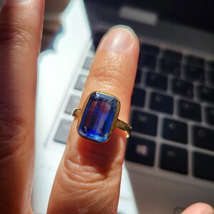 May include: A gold ring featuring a large, rectangular blue gemstone. The gemstone is set in a gold band with a simple, elegant design. The ring is shown on a finger, with a laptop keyboard in the background.
