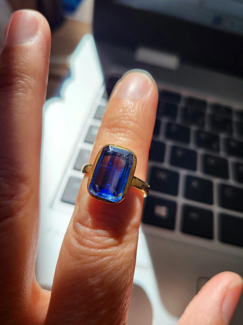 May include: A gold ring featuring a large, rectangular blue gemstone. The gemstone is set in a gold band with a simple, elegant design. The ring is shown on a finger, with a laptop keyboard in the background.