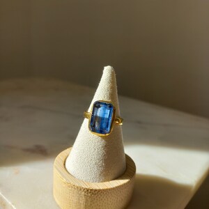 May include: A gold ring with a large, rectangular, blue gemstone. The ring is displayed on a white cone-shaped jewelry holder with a wooden base. The gemstone is a vibrant blue color, and the gold setting complements the stone.