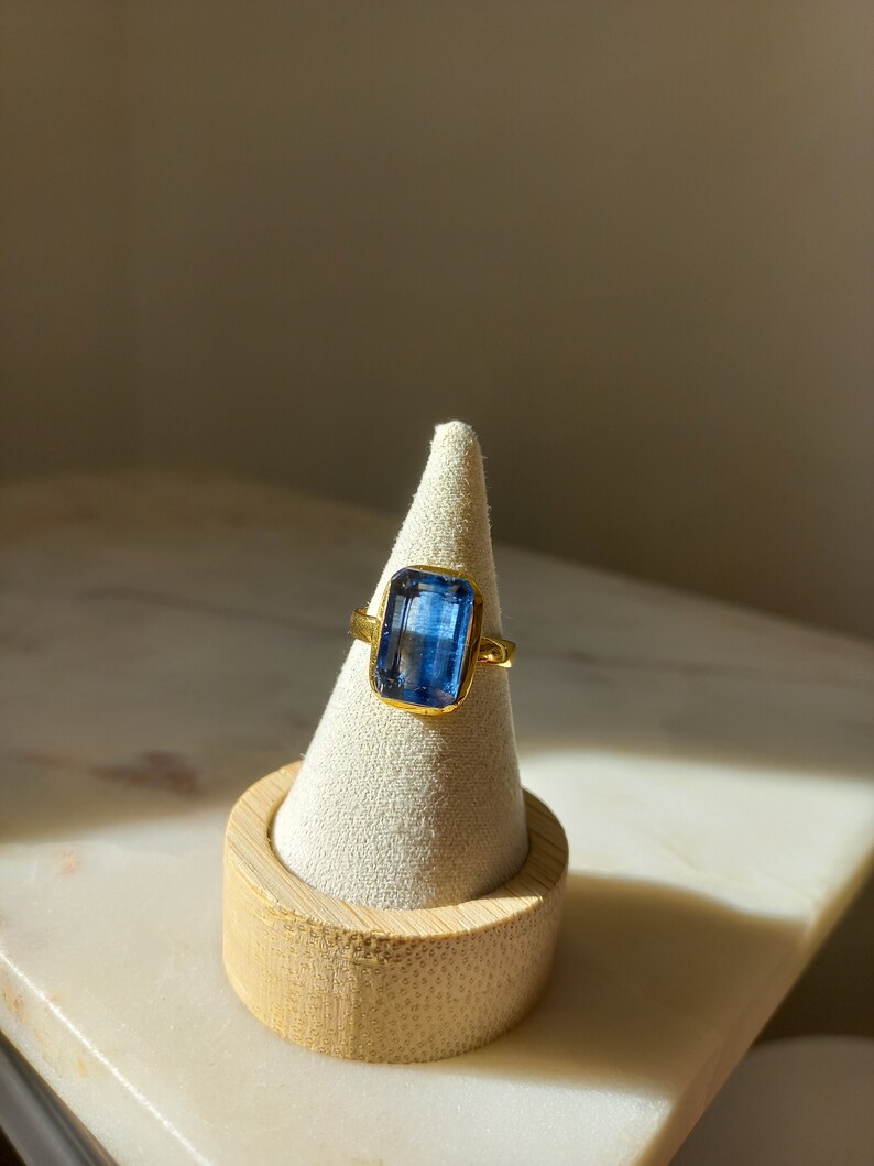 May include: A gold ring with a large, rectangular, blue gemstone. The ring is displayed on a white cone-shaped jewelry holder with a wooden base. The gemstone is a vibrant blue color, and the gold setting complements the stone.