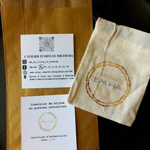 以下が含まれることがあります： 「L'ATELIER D'ARTS LE 768 EM-HA」というテキストと、ジュエリーメーカーの連絡先が書かれた白いカード。カードには、「Cr&eacute;atrice de bijoux en pierres naturelles」と「Certificat d'authenticit&eacute;」というテキストも含まれています。