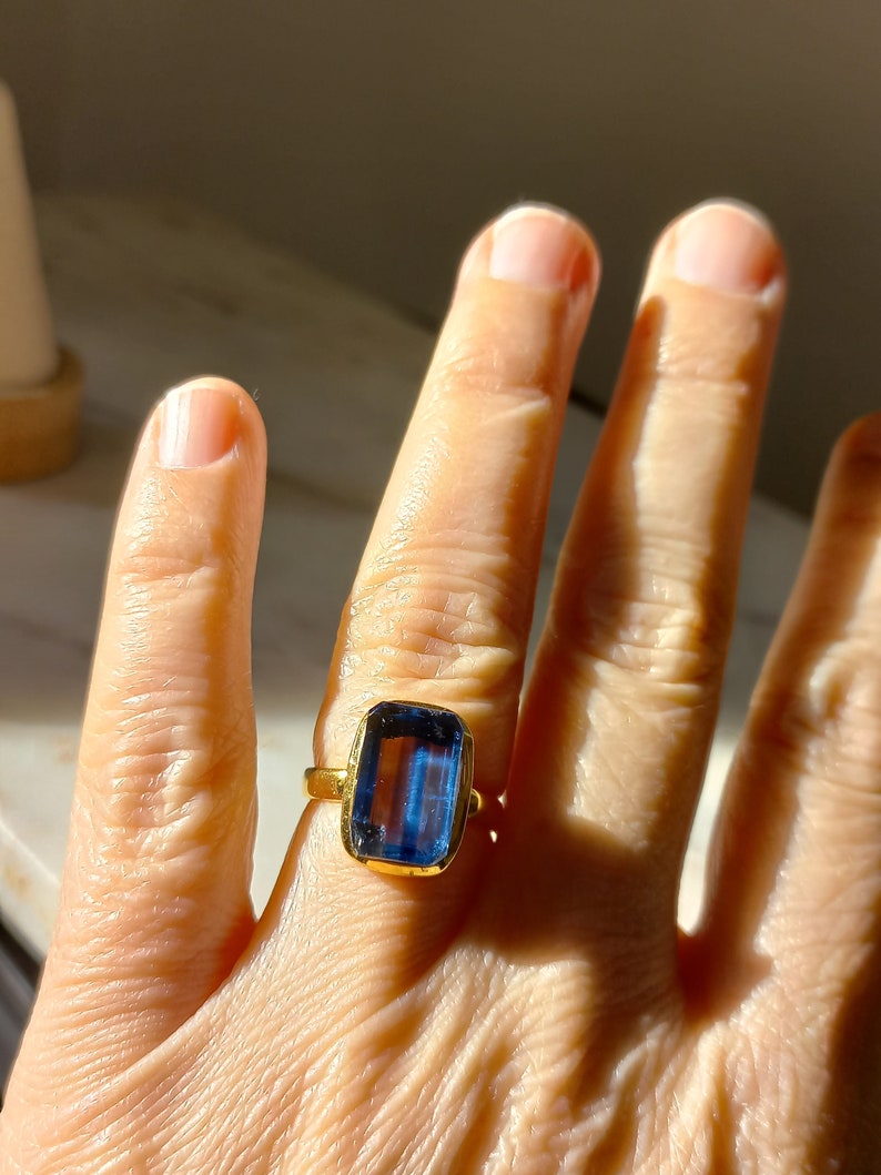 May include: A gold ring featuring a large, rectangular, blue gemstone. The ring's band is a simple, polished gold. The gemstone is a deep, vibrant blue, with a rectangular cut. The ring is worn on a finger.
