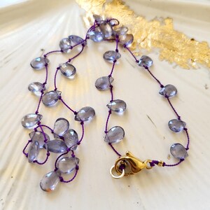 May include: A delicate necklace featuring teardrop-shaped, translucent blue-grey beads strung on a purple cord. The necklace is secured with a gold-colored clasp. The jewelry rests on a white, shell-shaped dish with gold leaf accents.