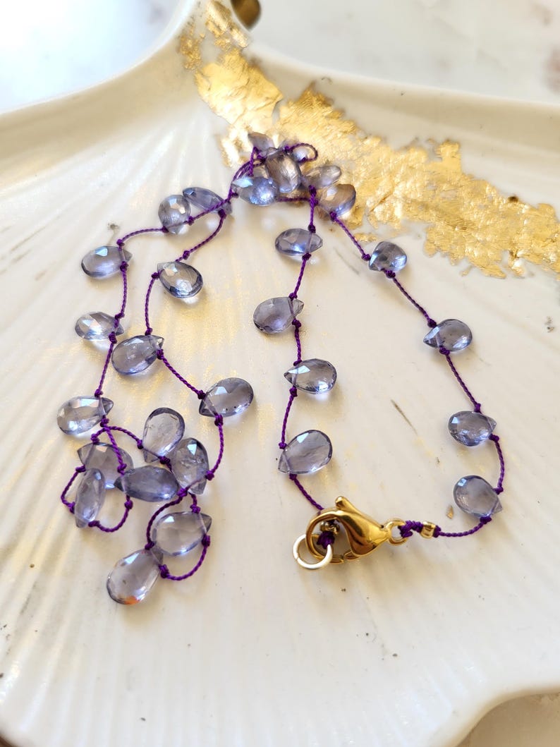 May include: A delicate necklace featuring teardrop-shaped, translucent blue-grey beads strung on a purple cord. The necklace is secured with a gold-colored clasp. The jewelry rests on a white, shell-shaped dish with gold leaf accents.