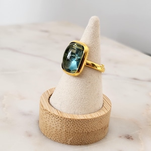 May include: A gold ring with a large, square, blue gemstone. The ring is displayed on a small, wooden ring holder.