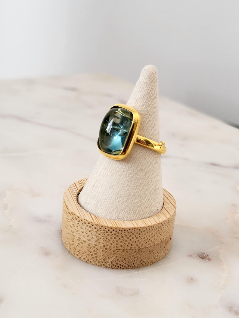 May include: A gold ring with a large, square, blue gemstone. The ring is displayed on a small, wooden ring holder.