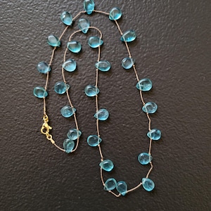 Pu&ograve; includere: Una delicata collana con pietre preziose blu chiaro a forma di goccia infilate su una sottile catena color oro. La collana ha una chiusura color oro. Le pietre sono uniformemente distanziate lungo la catena.
