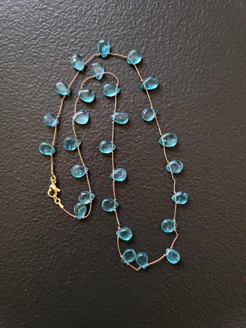 Pu&ograve; includere: Una delicata collana con pietre preziose blu chiaro a forma di goccia infilate su una sottile catena color oro. La collana ha una chiusura color oro. Le pietre sono uniformemente distanziate lungo la catena.