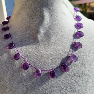May include: A necklace featuring a series of faceted, teardrop-shaped purple amethyst gemstones strung along a thin, dark purple thread. The stones are spaced evenly apart, creating a delicate, minimalist arrangement of natural violet crystals.