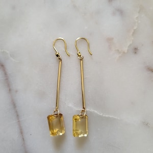 May include: A pair of gold drop earrings featuring a long, slender gold bar suspended from a hook wire, with a rectangular, faceted yellow gemstone dangling at the base of each earring. The design is symmetrical, showcasing polished metal and clear stones.