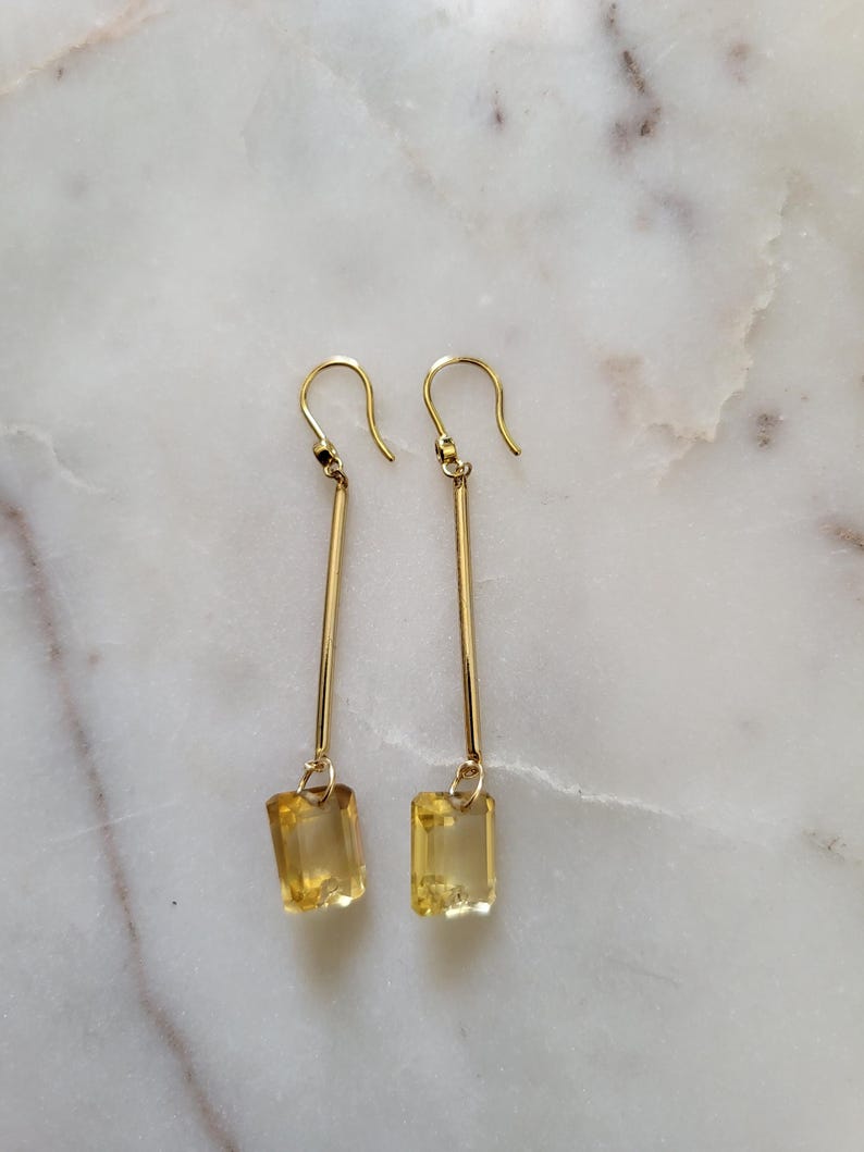 May include: A pair of gold drop earrings featuring a long, slender gold bar suspended from a hook wire, with a rectangular, faceted yellow gemstone dangling at the base of each earring. The design is symmetrical, showcasing polished metal and clear stones.