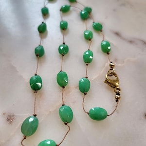 May include: A delicate necklace featuring faceted, oval-shaped green beads strung on a thin, brown cord. The necklace has a gold-colored clasp. The beads are a vibrant, light green color.