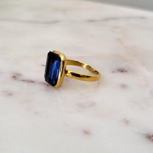 May include: A gold-toned ring featuring a large, rectangular, deep blue gemstone. The gemstone is set in a gold bezel, and the band is a simple, polished gold. The ring is displayed on a white and gray marble surface.