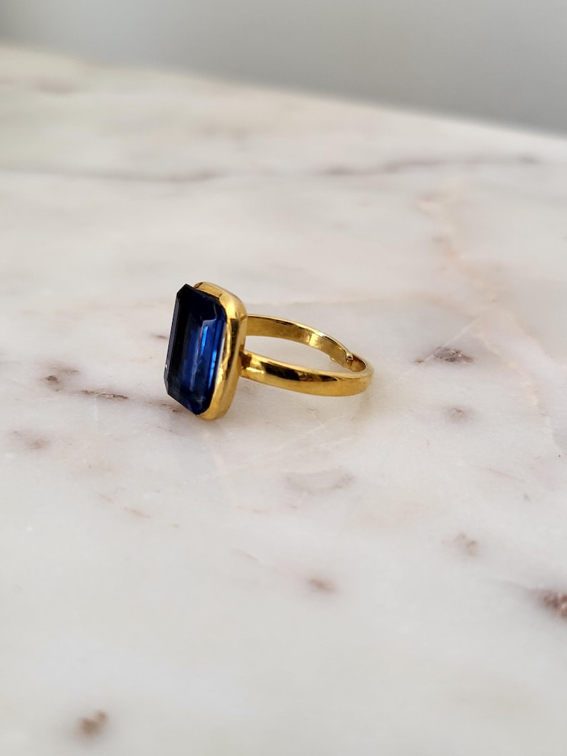 May include: A gold-toned ring featuring a large, rectangular, deep blue gemstone. The gemstone is set in a gold bezel, and the band is a simple, polished gold. The ring is displayed on a white and gray marble surface.
