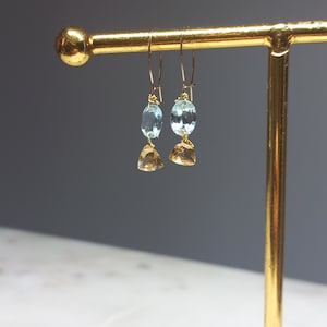 Op de afbeelding: Goudkleurige oorbellen met een lichtblauwe ovale edelsteen en een geelbruine driehoekige edelsteen, hangend aan een gouden draad. De oorbellen worden gepresenteerd op een gouden sieradenstandaard tegen een grijze achtergrond.