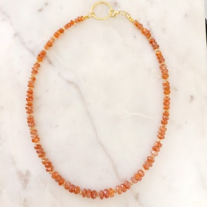 Peut inclure: Un collier compos&eacute; de petites pierres pr&eacute;cieuses orange-rouge &agrave; facettes, enfil&eacute;es sur un fil dor&eacute;. Il est dot&eacute; d'un fermoir dor&eacute; et d'un pendentif circulaire. Les pierres sont espac&eacute;es de mani&egrave;re uniforme, cr&eacute;ant un design d&eacute;licat et &eacute;l&eacute;gant.