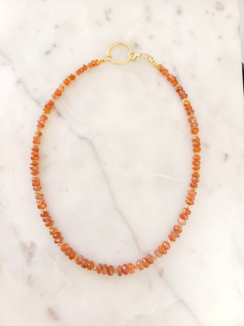 Peut inclure: Un collier compos&eacute; de petites pierres pr&eacute;cieuses orange-rouge &agrave; facettes, enfil&eacute;es sur un fil dor&eacute;. Il est dot&eacute; d'un fermoir dor&eacute; et d'un pendentif circulaire. Les pierres sont espac&eacute;es de mani&egrave;re uniforme, cr&eacute;ant un design d&eacute;licat et &eacute;l&eacute;gant.