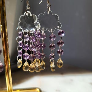 May include: Cloud-shaped earrings made of clear material, with strands of purple and yellow beads hanging down. The earrings have silver hooks and are displayed on a gold stand. The beads are arranged to resemble raindrops.
