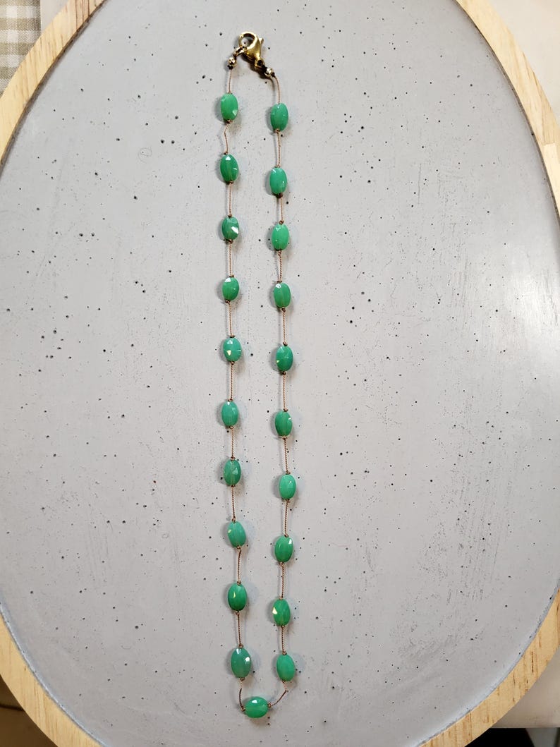 May include: A delicate necklace featuring oval-shaped, light green beads strung on a thin, gold-colored chain. The beads are evenly spaced, creating a simple yet elegant design. The necklace is displayed on a light gray surface.