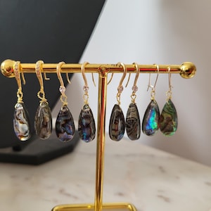 May include: A collection of teardrop-shaped earrings with iridescent abalone shells, displayed on a gold-colored earring stand. The earrings have gold-colored hooks and are arranged in a row, showcasing their unique colors and patterns.