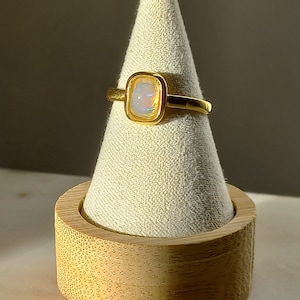 Peut inclure: Une bague en or avec une opale blanche sertie dans un bézel carré. La bague est présentée sur un porte-bague en bois.