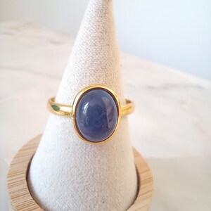 K&ouml;nnte beinhalten: Ein goldener Ring mit einem blauen ovalen Edelstein. Der Ring wird auf einem wei&szlig;en, kegelf&ouml;rmigen Ringst&auml;nder pr&auml;sentiert.