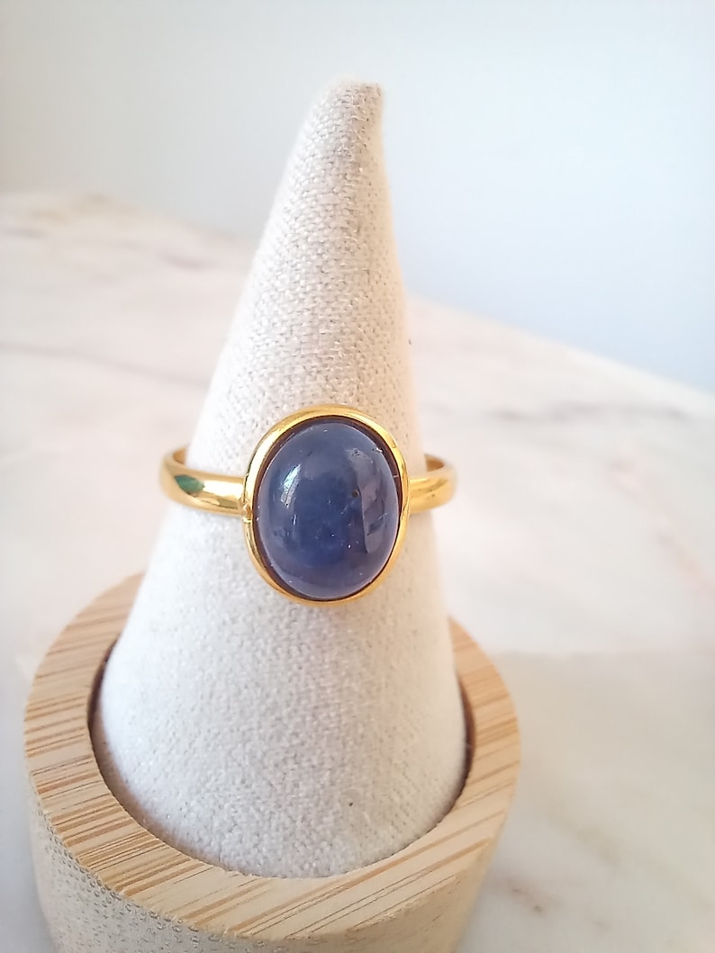 K&ouml;nnte beinhalten: Ein goldener Ring mit einem blauen ovalen Edelstein. Der Ring wird auf einem wei&szlig;en, kegelf&ouml;rmigen Ringst&auml;nder pr&auml;sentiert.