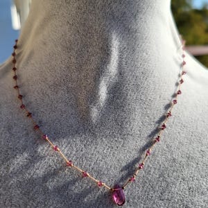 May include: A delicate necklace featuring small, round, pink gemstones strung on a thin, gold-colored wire. A larger, teardrop-shaped pink gemstone hangs as a pendant. The necklace is displayed on a gray surface.