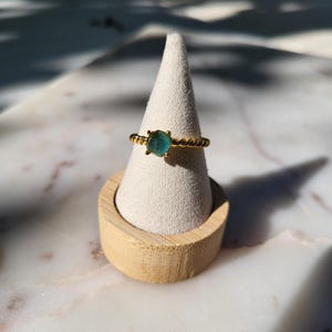 May include: A gold ring with a square, teal-colored gemstone. The ring has a twisted band design. The ring is displayed on a white cone-shaped jewelry holder, which is set on a wooden base.