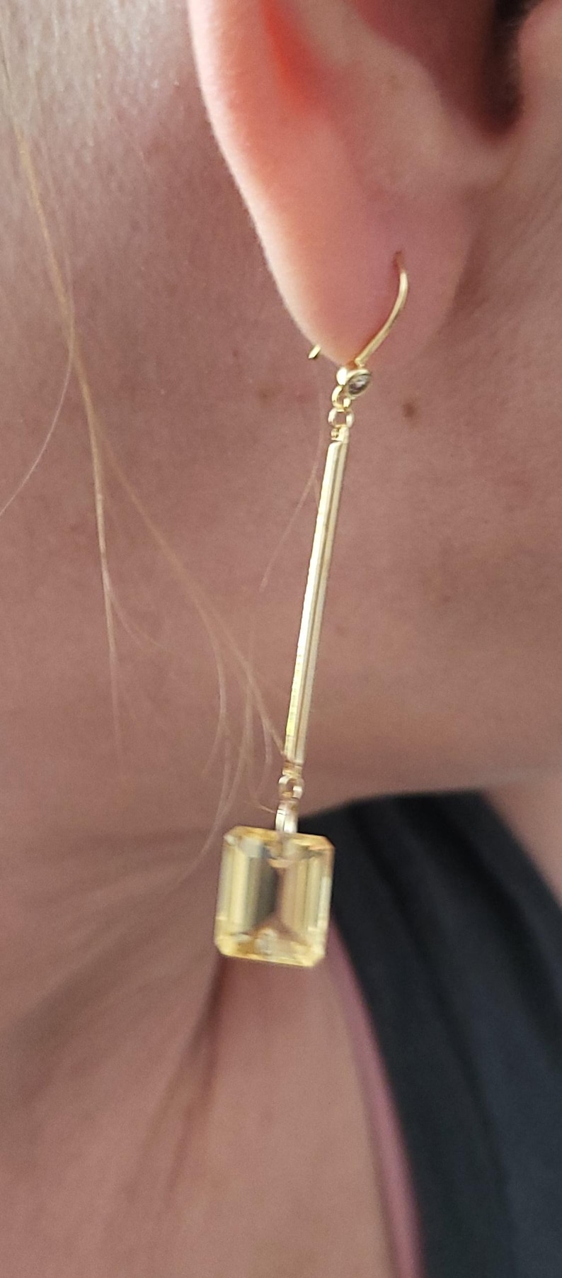 May include: A long gold drop earring featuring a thin, vertical gold bar suspended from a hook. At the base of the bar hangs a single, rectangular, light yellow gemstone with a faceted cut, creating a clean and elongated silhouette.