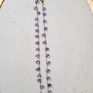 May include: A delicate necklace featuring teardrop-shaped, translucent blue-gray beads strung on a thin purple cord. The necklace has a gold-colored clasp and is displayed on a light gray surface.