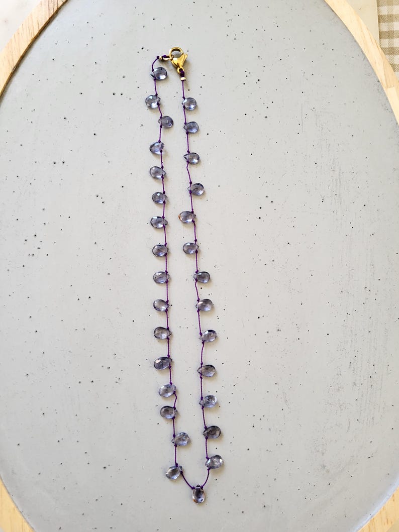 May include: A delicate necklace featuring teardrop-shaped, translucent blue-gray beads strung on a thin purple cord. The necklace has a gold-colored clasp and is displayed on a light gray surface.