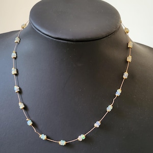 May include: Delicate necklace featuring small, iridescent opal stones strung on a fine gold-colored chain. The opals display a range of colors, including white, blue, and green. The necklace is displayed on a black mannequin.